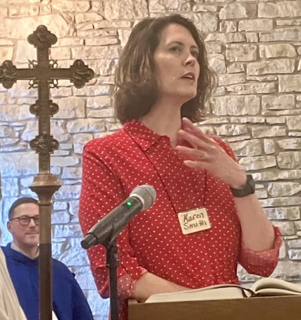 Karen Smith speaking at St. Thomas Episcopal Church podium with cross and stone wall backdrop.
